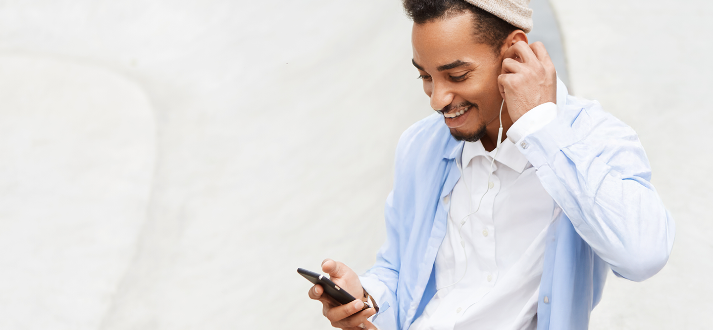 Homem jovem com barba e gorro bege, vestindo camisa branca e sobreposição azul clara. Ele sorri enquanto olha para o celular e ajusta fones de ouvido brancos.