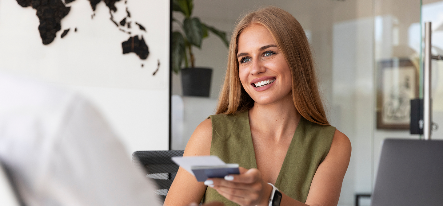 Mulher loira sorridente vestindo blusa verde oliva, segurando documentos em um escritório com um mapa-múndi ao fundo.