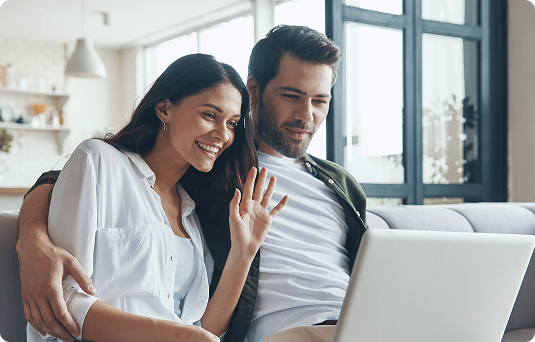 Casal jovem sentado no sofá em videochamada pelo notebook, com a mulher de blusa branca acenando e sorrindo e o homem de camiseta branca ao lado, em ambiente doméstico iluminado.