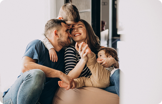 Um casal e duas crianças pequenas sentados no chão de uma sala iluminada. O pai olha para a mãe enquanto ela sorri, abraçada às crianças. O clima é de carinho e união familiar.
