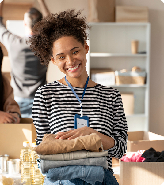 Mulher jovem negra sorridente, com cabelo cacheado preso, vestindo blusa listrada em preto e branco e um crachá azul de "Volunteer". Ela segura uma pilha de roupas dobradas em um centro de doações.