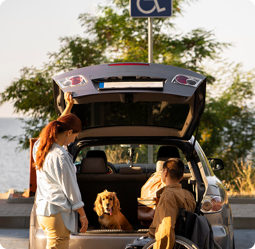 Casal ao lado de carro com porta-malas aberto e cão da raça cocker spaniel sentado dentro, estacionado em vaga para pessoas com deficiência, com árvores e água ao fundo.