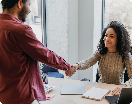 Mulher jovem de cabelo cacheado e suéter bege sorrindo enquanto aperta a mão de um homem de camisa vinho sobre mesa de escritório com notebook e caderno.