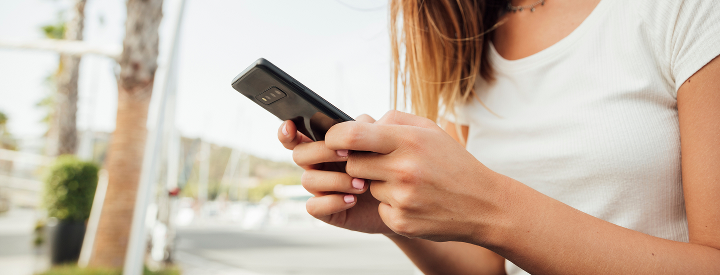 Mulher jovem segurando e digitando em um smartphone preto com as duas mãos, ao ar livre em ambiente urbano ensolarado.