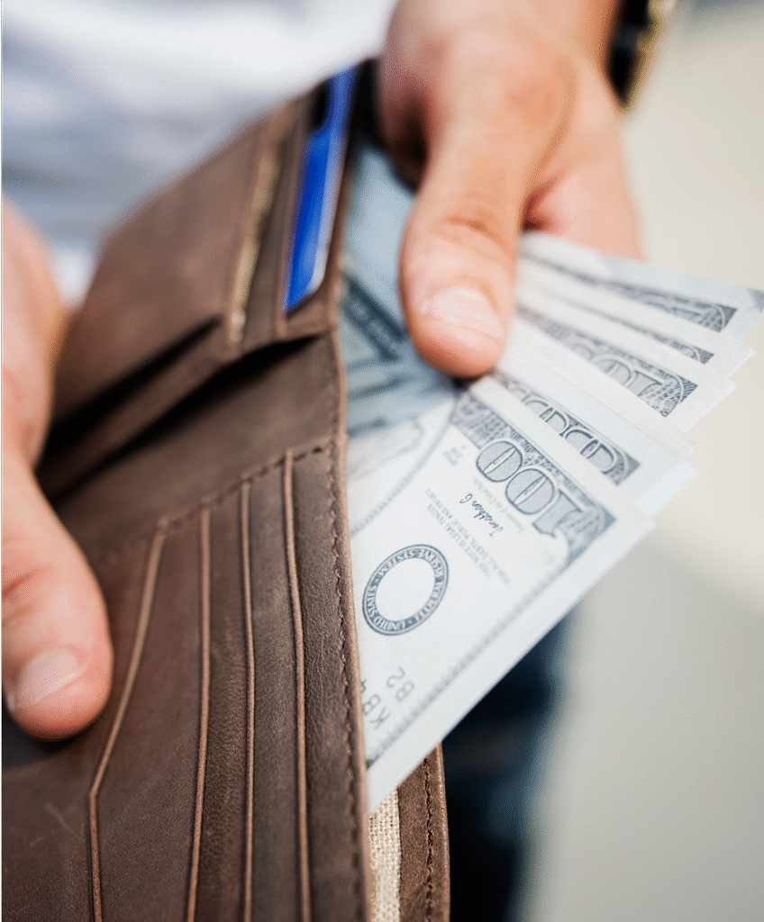 Mãos segurando uma carteira de couro marrom aberta com notas de dólar sendo retiradas e um cartão azul visível no compartimento interno.