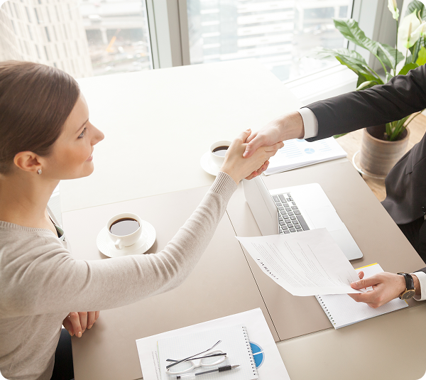 Vista aérea de duas pessoas trocando aperto de mão sobre mesa de reunião com documentos, laptop e xícara de café.