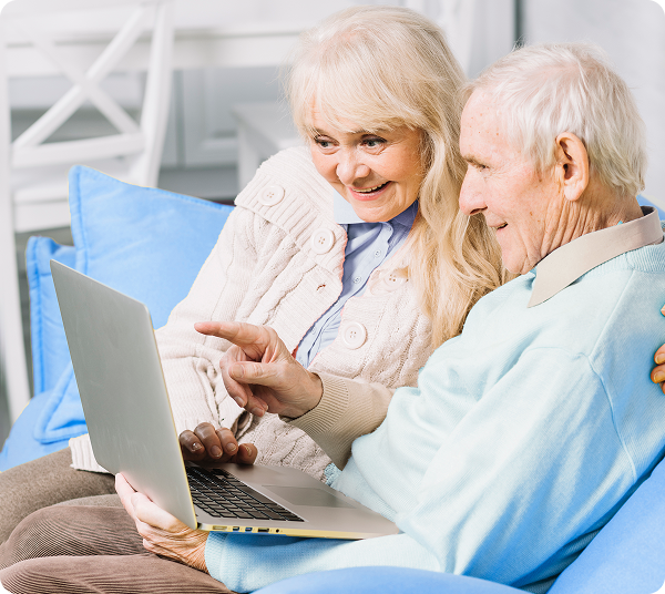Casal de idosos sentado no sofá sorrindo enquanto usa laptop juntos, com a mulher apontando para a tela.