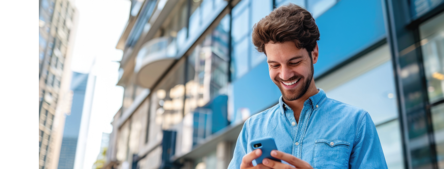 Homem jovem de camisa jeans sorrindo enquanto usa smartphone em área urbana com prédios modernos ao fundo.