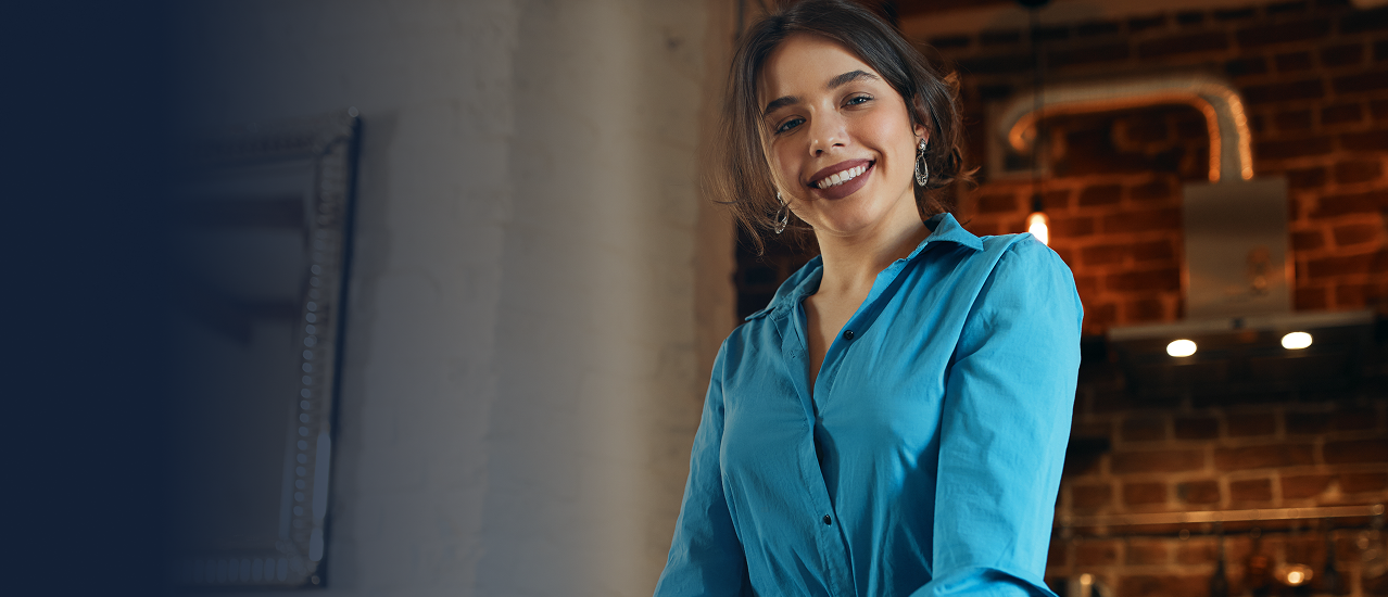 Mulher jovem de camisa azul e cabelos semi-presos sorrindo em ambiente com parede de tijolos ao fundo.