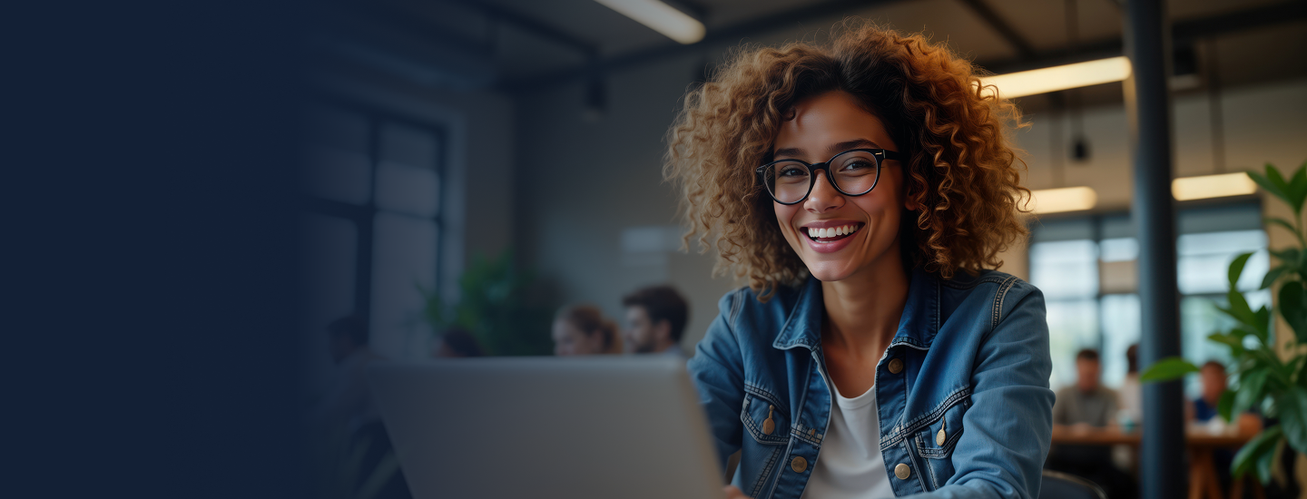 Mulher jovem de cabelos cacheados, óculos e jaqueta jeans sorrindo em frente ao laptop em espaço de coworking.