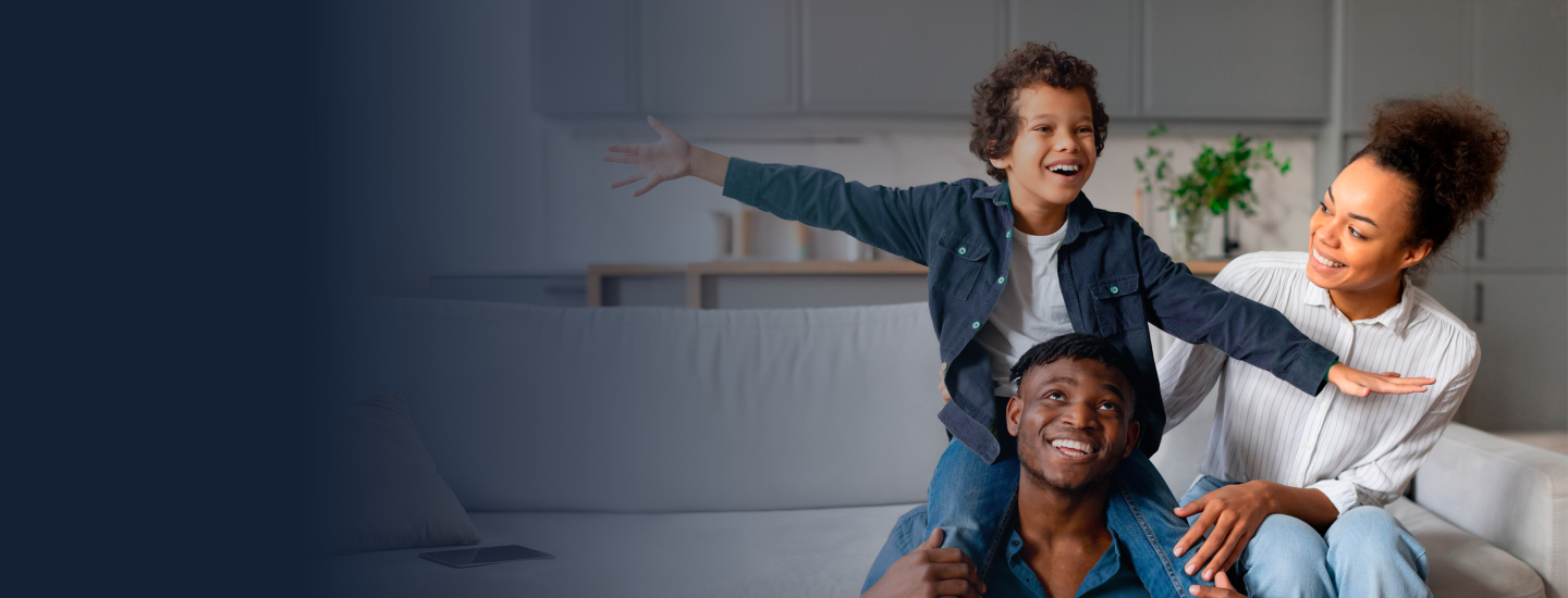 Família jovem brincando na sala de casa, com filho sentado nos ombros do pai e braços abertos enquanto a mãe sorri ao lado.