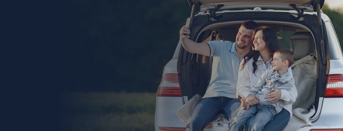 Família composta por casal e filho pequeno sentados no porta-malas de um carro tirando uma selfie ao ar livre.