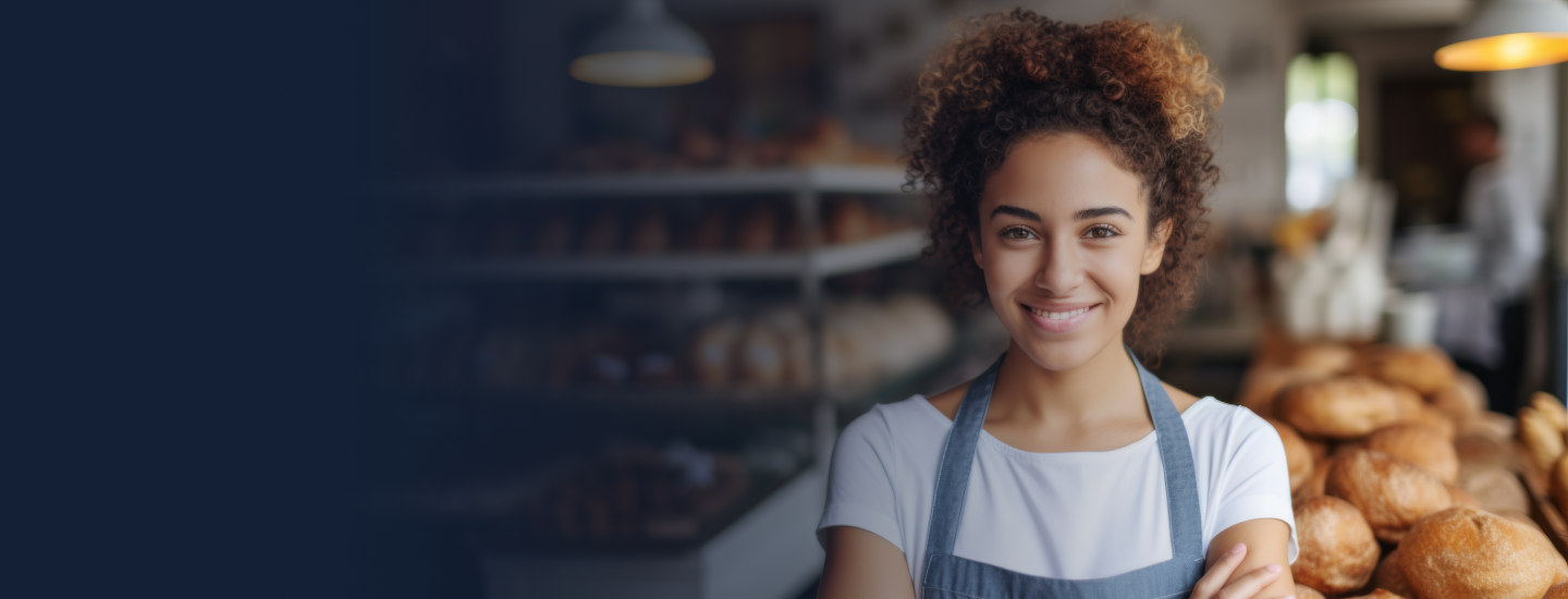 Jovem padeira de avental sorrindo com os braços cruzados em frente a pães frescos em padaria.