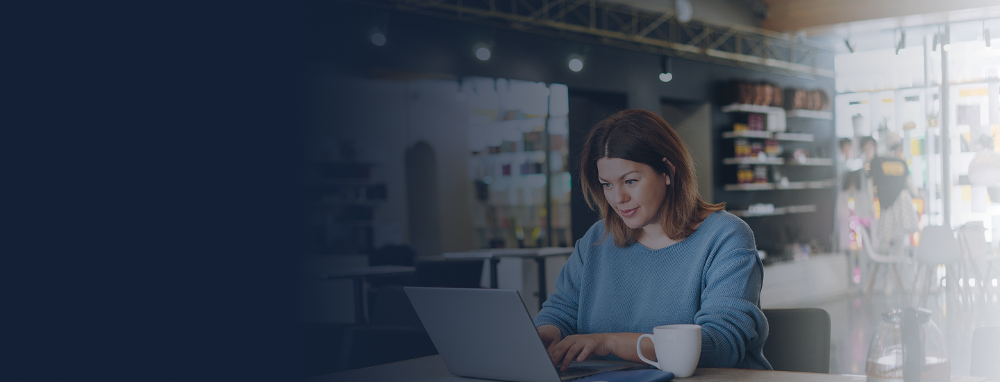 Mulher adulta trabalhando no laptop em café ou espaço de coworking com xícara ao lado.