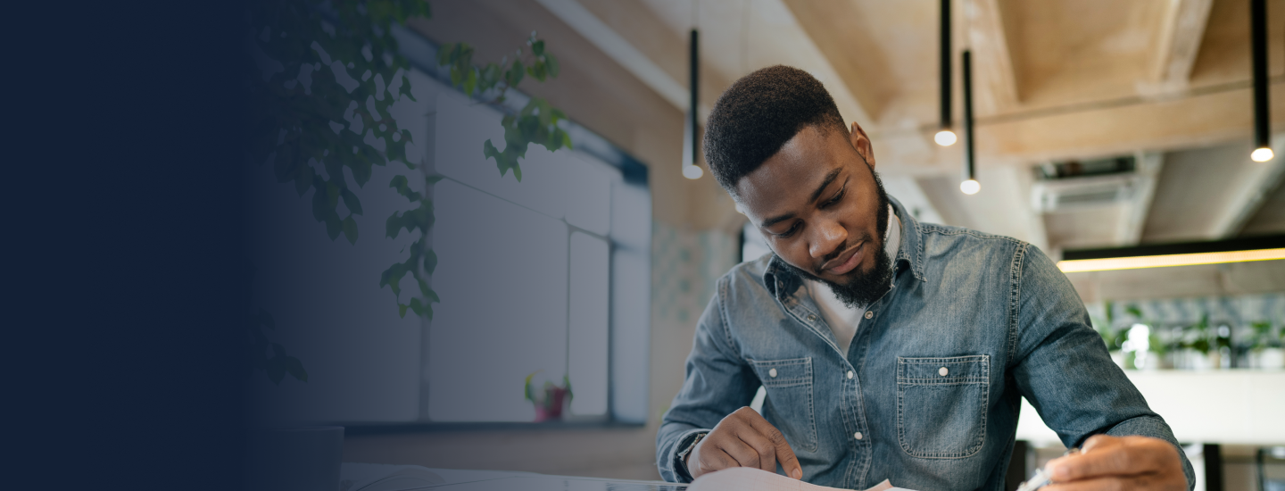 Homem jovem de camisa jeans escrevendo em documento sobre mesa em espaço de trabalho moderno.