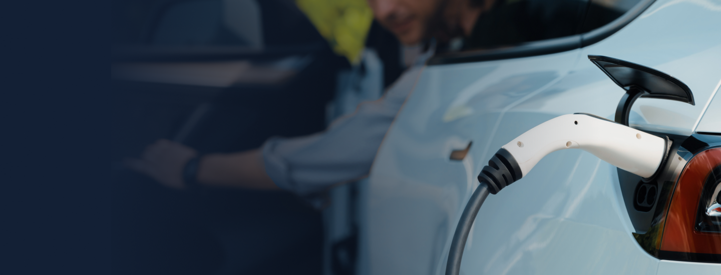 Cabo de carregamento conectado a um carro elétrico com pessoa ao fundo segurando um tablet.