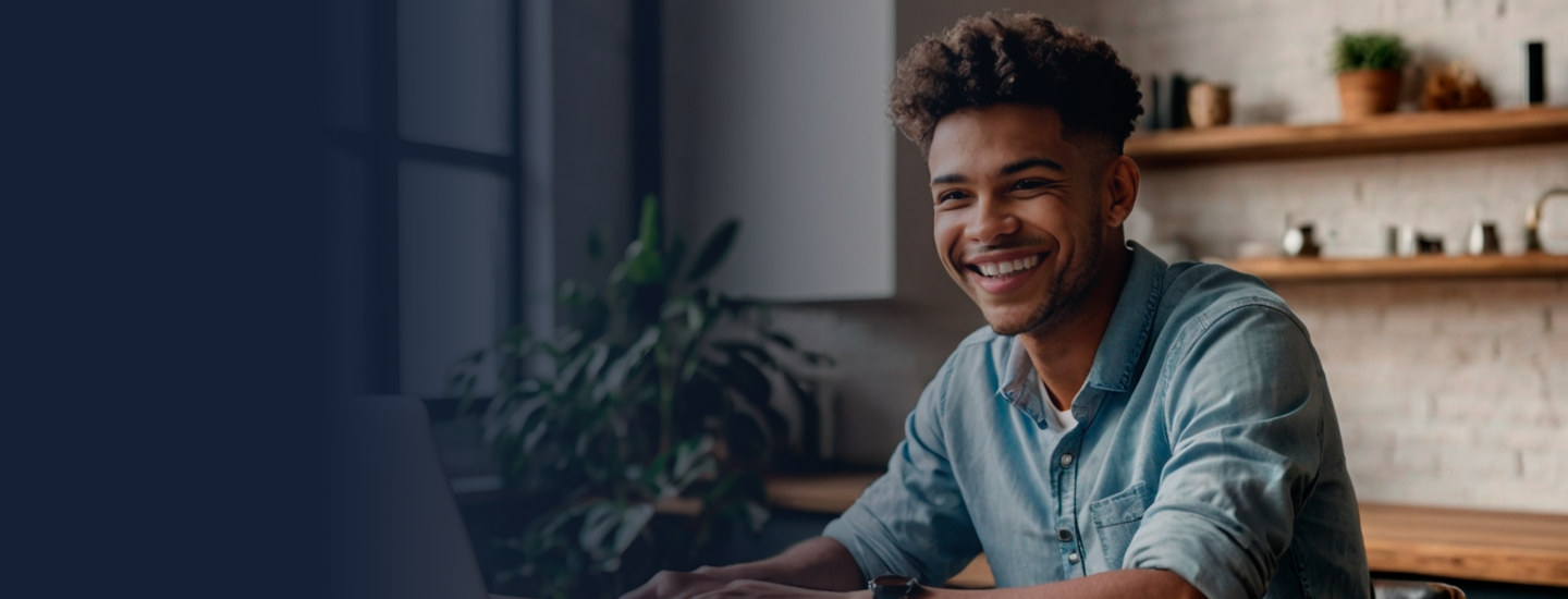 Homem jovem de cabelos cacheados e camisa jeans sorrindo em frente ao laptop em ambiente doméstico aconchegante.