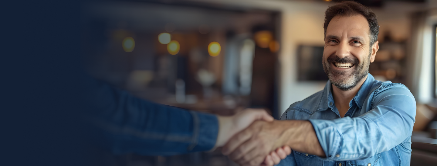 Homem adulto de camisa jeans sorrindo enquanto cumprimenta alguém com aperto de mão em ambiente interno aquecido.