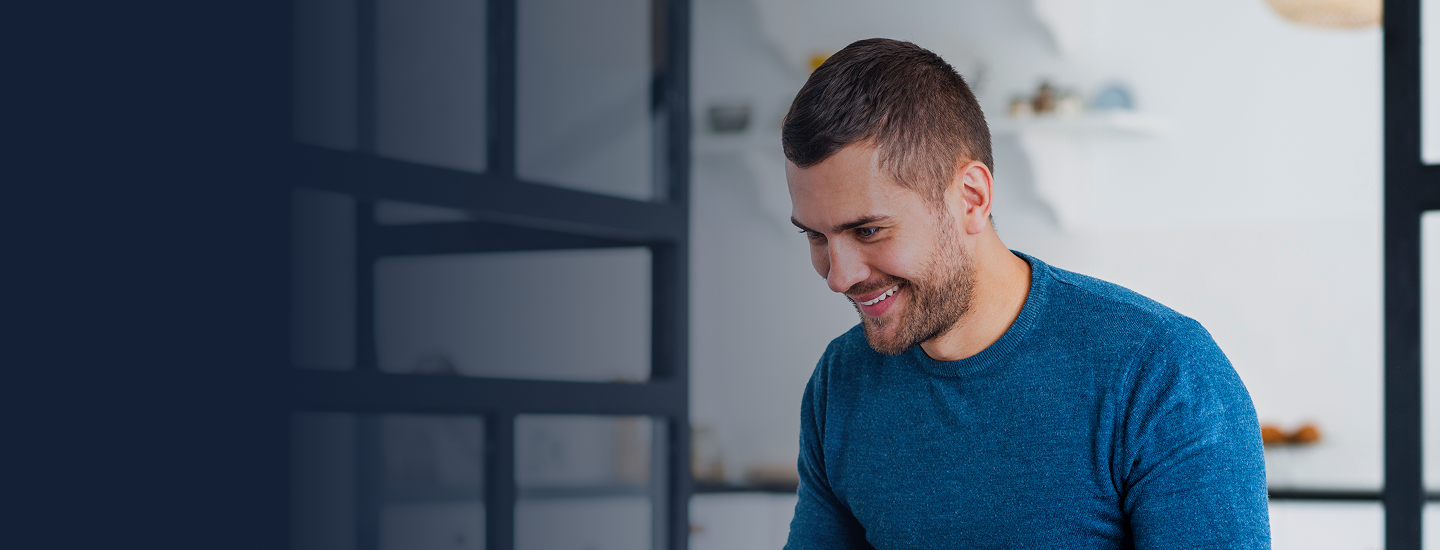 Homem jovem de suéter azul sorrindo enquanto olha para baixo em ambiente moderno com divisórias de vidro.