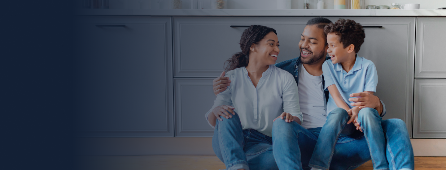 Família jovem composta por casal e filho sentados no chão da cozinha sorrindo e interagindo entre si.