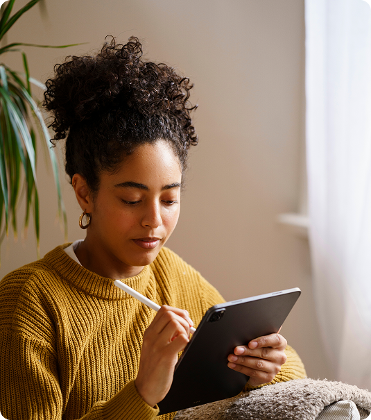 Mulher jovem de cabelos cacheados presos escrevendo em tablet com caneta stylus em ambiente doméstico.