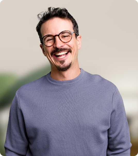 Homem adulto de óculos e moletom cinza sorrindo em frente a fundo neutro.