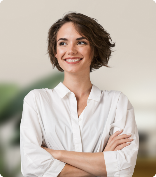 Mulher jovem de cabelos curtos e camisa branca sorrindo com os braços cruzados em fundo neutro.