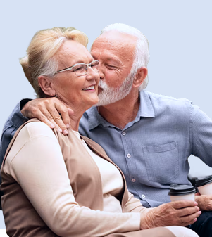 Casal de idosos em momento de afeto, com o homem dando um beijo na bochecha da mulher em fundo azul claro.
