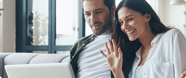 Casal jovem em videochamada pelo laptop na sala de estar, com a mulher acenando para a tela.