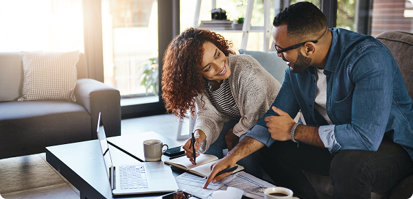 Casal jovem sorrindo enquanto analisa documentos e usa laptop em sala de estar.