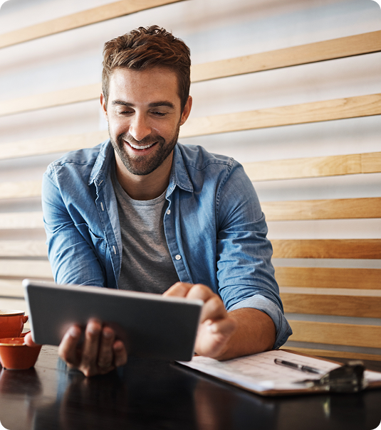 Homem jovem sorrindo enquanto usa um tablet em uma mesa com caderno e xícara.