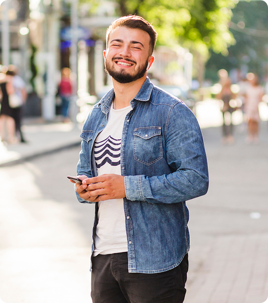 Homem jovem sorrindo e segurando smartphone em calçada arborizada ao ar livre.