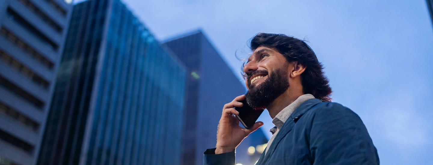 Homem jovem sorrindo enquanto fala ao telefone em área urbana com prédios ao fundo.
