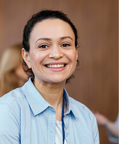 Mulher adulta sorrindo, vestindo camisa azul clara e crachá, em ambiente interno.