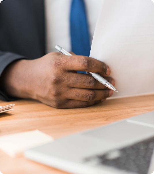 Mão de homem segurando uma caneta e documentos sobre uma mesa de escritório.