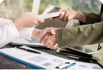 Duas pessoas apertando as mãos sobre uma mesa com gráficos e documentos, enquanto uma delas entrega um cartão.